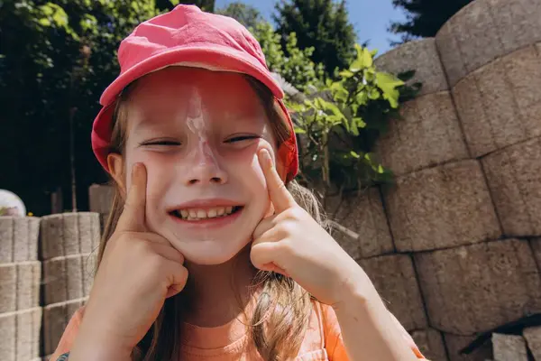 Pembe şapkalı genç bir kız güneşli bir yaz gününde yüzüne güneş kremi sürer. Cilt bakımı, güneş koruması ve çocuk sağlığı konsepti. Yüksek kalite fotoğraf