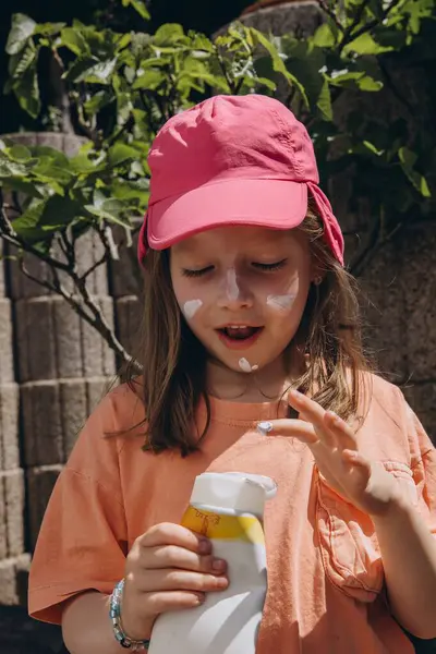 Pembe şapkalı genç bir kız güneşli bir yaz gününde yüzüne güneş kremi sürer. Cilt bakımı, güneş koruması ve çocuk sağlığı konsepti. Yüksek kalite fotoğraf