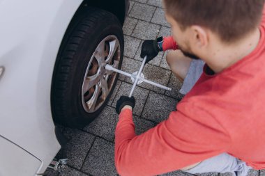 Evinin önünde lastik değiştiren bir adamın yakın çekimi. Araç kriko ile kaldırılır, aletler yakınlarda durur. Olay yeri, gündüz vakti elle tamir edilen bir anı yakalıyor. Yüksek kalite fotoğraf