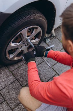 Evinin önünde lastik değiştiren bir adamın yakın çekimi. Araç kriko ile kaldırılır, aletler yakınlarda durur. Olay yeri, gündüz vakti elle tamir edilen bir anı yakalıyor. Yüksek kalite fotoğraf