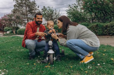 Parkta iki yaşındaki oğulları olan bir anne ve baba. Bebek küçük bir bisiklete biniyor, dışarıda eğleniyor, eğleniyor ve öğreniyor. Yüksek kalite fotoğraf