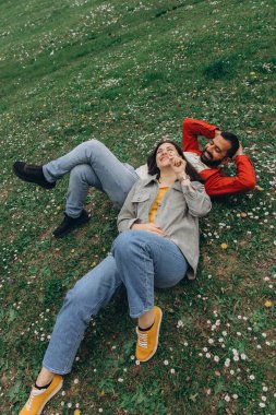 İki insan, kadın ve erkek yazın çimenlikte küçük kır çiçekleriyle birlikte dinleniyor, dışarıda huzurlu bir anın tadını çıkarıyorlar. Günlük giysiler ve doğal çevre sakin ve otantik bir ortam yaratıyor. Yüksek kalite fotoğraf