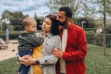 Genç bir aile parkın dışında birlikte vakit geçiriyor. Anne kucağında bir bebekle dolaşırken, baba onları kucaklıyor, günlük giysiler içinde, doğada huzurlu bir günün tadını çıkarıyor. Yüksek kalite fotoğraf