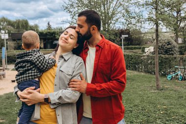 Genç bir aile parkın dışında birlikte vakit geçiriyor. Anne kucağında bir bebekle dolaşırken, baba onları kucaklıyor, günlük giysiler içinde, doğada huzurlu bir günün tadını çıkarıyor. Yüksek kalite fotoğraf