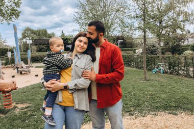 Genç bir aile parkın dışında birlikte vakit geçiriyor. Anne kucağında bir bebekle dolaşırken, baba onları kucaklıyor, günlük giysiler içinde, doğada huzurlu bir günün tadını çıkarıyor. Yüksek kalite fotoğraf