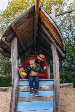 Baba ve küçük çocuk açık hava oyun parkında tahta bir oyun evinde birlikte oynuyorlar. Çocuk, doğal ahşap dokular ve yeşilliklerle çevrili sarı bir kepçeden kum döküyor. Yüksek kalite fotoğraf
