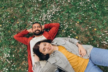 İki insan, kadın ve erkek yazın çimenlikte küçük kır çiçekleriyle birlikte dinleniyor, dışarıda huzurlu bir anın tadını çıkarıyorlar. Günlük giysiler ve doğal çevre sakin ve otantik bir ortam yaratıyor. Yüksek kalite fotoğraf