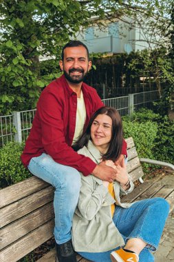 Mutlu çift dışarıda bir bankta oturuyor, gülümsüyor ve birbirlerine sarılıyorlar. Neşeli, sevecen bir an. Sevgiyi, bağlantıyı ve tasasız birlikteliği doğal ortamında yakalıyor. Yüksek kalite fotoğraf