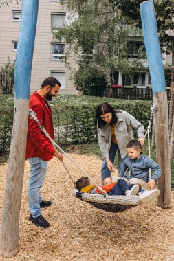 İki oğlu olan mutlu bir aile oyun parkında büyük bir salıncakta birlikte oynuyorlar. Aileler gülümsüyor, bağ kuruyor ve çocuklarla dışarıda kaliteli zaman geçiriyorlar. Ebeveynlik ve çocukluk kavramı. Yüksek kalite fotoğraf