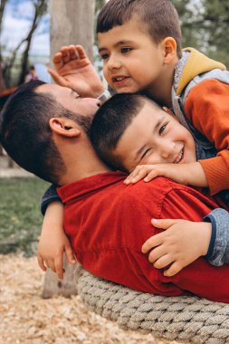 Baba iki oğlunu dışarıda kucaklıyor. Bir çocuk sıkıca sarılırken gülümser, diğeri yukarıdan sarılır. Sevgi ve bağlılık dolu sıcak bir aile anı. Yüksek kalite fotoğraf