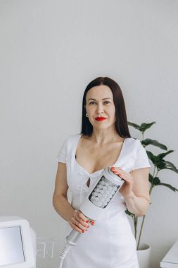 Kendine güvenen beyaz üniformalı kadın kozmetik uzmanı modern bir güzellik kliniğinde endosfer terapi cihazı tutuyor. Yüz ve vücut yenilenmesi için zararsız bir tedavi. Yüksek kalite fotoğraf