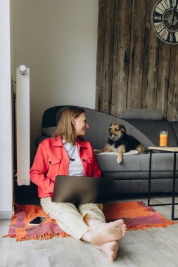Evde dizüstü bilgisayarla uzaktan çalışan bir kadın, kanepede yatan köpeğine gülümsüyor. Sıcak atmosfer, iş hayatı dengesi ve evcil hayvanlarla duygusal bağ. Yüksek kalite fotoğraf