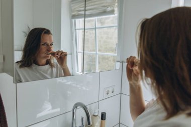 Parlak banyoda dişlerini fırçalayan bir kadın. Sabah rutini, diş bakımı, hijyen, tazelik, sağlık, kişisel bakım, doğal güzellik, ağız temizliği, sağlıklı yaşam tarzı, günlük ritüel, sağlık. Yüksek kalite fotoğraf