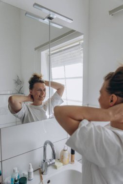 Aynanın önünde saç yapan kadın. Sabah rutini, günlük görünüş, güzellik, sağIık, sağlıklı cilt, bencillik, taze başlangıç, ev banyosu, tımar, hijyen, ritüel, mutlu ruh hali, gerçek hayat. Yüksek kalite fotoğraf