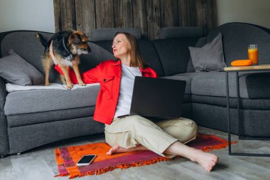 Evde dizüstü bilgisayarla uzaktan çalışan bir kadın, kanepede yatan köpeğine gülümsüyor. Sıcak atmosfer, iş hayatı dengesi ve evcil hayvanlarla duygusal bağ. Yüksek kalite fotoğraf