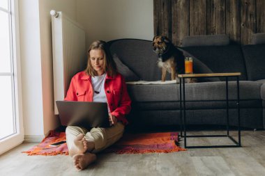Evde dizüstü bilgisayarla çalışan serbest çalışan bir kadın, rahat oturma odasında yerde oturuyor, yalınayak ve rahatlamış, köpeğini okşuyor. Uzaktan çalışma ve hayvan dostu yaşam tarzı konsepti. Yüksek kalite fotoğraf