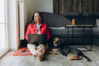 Evde dizüstü bilgisayarla çalışan serbest çalışan bir kadın, rahat oturma odasında yerde oturuyor, yalınayak ve rahatlamış, köpeğini okşuyor. Uzaktan çalışma ve hayvan dostu yaşam tarzı konsepti. Yüksek kalite fotoğraf
