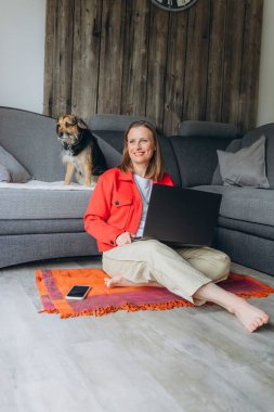 Evde dizüstü bilgisayarla çalışan serbest çalışan bir kadın, rahat oturma odasında yerde oturuyor, yalınayak ve rahatlamış, köpeğini okşuyor. Uzaktan çalışma ve hayvan dostu yaşam tarzı konsepti. Yüksek kalite fotoğraf