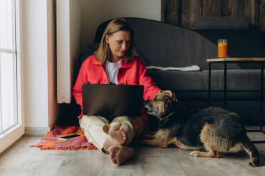Evde dizüstü bilgisayarla çalışan serbest çalışan bir kadın, rahat oturma odasında yerde oturuyor, yalınayak ve rahatlamış, köpeğini okşuyor. Uzaktan çalışma ve hayvan dostu yaşam tarzı konsepti. Yüksek kalite fotoğraf