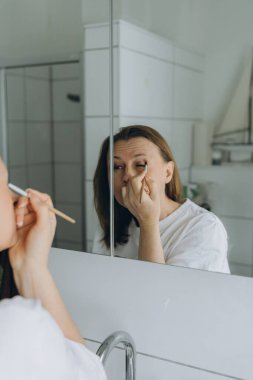 Orta yaşlı bir kadın banyo aynasının önünde fırçayla göz makyajı yapıyor. Günlük ev ortamında güzellik rutini ve kişisel bakım. Yüksek kalite fotoğraf