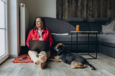 Evde dizüstü bilgisayarla çalışan serbest çalışan bir kadın, rahat oturma odasında yerde oturuyor, yalınayak ve rahatlamış, köpeğini okşuyor. Uzaktan çalışma ve hayvan dostu yaşam tarzı konsepti. Yüksek kalite fotoğraf