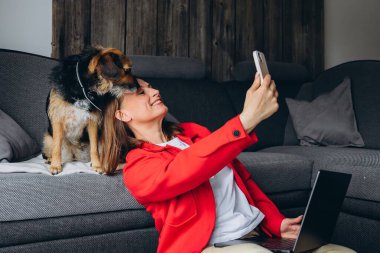 Bilgisayarla uzaktan çalışan bir kadın, evde köpeğiyle selfie çekiyor. Mutlu serbest çalışanlar evcil hayvanlarla eğleniyor, rahat yaşam alanında yerde oturuyor, gündelik dijital yaşam tarzı. Yüksek kalite fotoğraf
