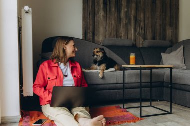 Evde dizüstü bilgisayarla uzaktan çalışan bir kadın, kanepede yatan köpeğine gülümsüyor. Sıcak atmosfer, iş hayatı dengesi ve evcil hayvanlarla duygusal bağ. Yüksek kalite fotoğraf