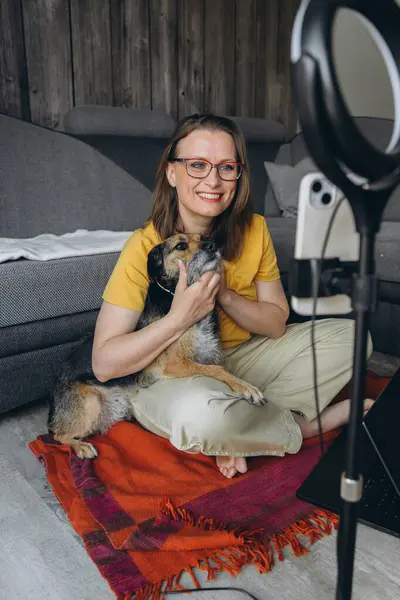 Gözlüklü gülümseyen kadın evde video kaydı yaparken köpeğini tutuyor. Samimi iç mekan, dizüstü bilgisayar ve çerçeve ışığı. Uzaktan çalışma ya da online danışma kurulumu. Yüksek kalite fotoğraf