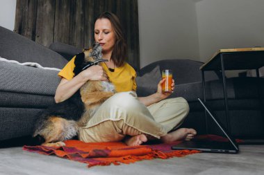 Uzaktan bilgisayarla çalışan, elinde meyve suyu olan, yerde köpeğiyle oturan gülümseyen bir kadın. Serbest çalışma anlayışı, sağlıklı yaşam tarzı ve evcil hayvanla iletişim. Yüksek kalite fotoğraf