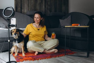 Uzaktan bilgisayarla çalışan, elinde meyve suyu olan, yerde köpeğiyle oturan gülümseyen bir kadın. Serbest çalışma anlayışı, sağlıklı yaşam tarzı ve evcil hayvanla iletişim. Yüksek kalite fotoğraf