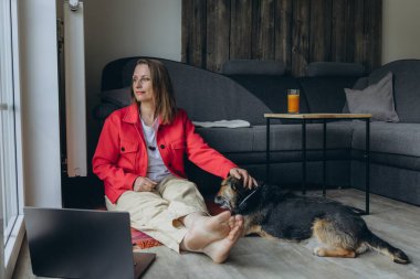 Evde dizüstü bilgisayarla çalışan serbest çalışan bir kadın, rahat oturma odasında yerde oturuyor, yalınayak ve rahatlamış, köpeğini okşuyor. Uzaktan çalışma ve hayvan dostu yaşam tarzı konsepti. Yüksek kalite fotoğraf