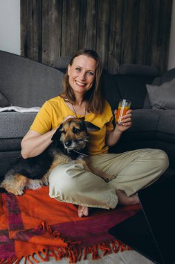 Uzaktan bilgisayarla çalışan, elinde meyve suyu olan, yerde köpeğiyle oturan gülümseyen bir kadın. Serbest çalışma anlayışı, sağlıklı yaşam tarzı ve evcil hayvanla iletişim. Yüksek kalite fotoğraf