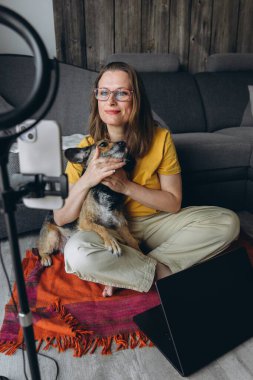 Gözlüklü gülümseyen kadın evde video kaydı yaparken köpeğini tutuyor. Samimi iç mekan, dizüstü bilgisayar ve çerçeve ışığı. Uzaktan çalışma ya da online danışma kurulumu. Yüksek kalite fotoğraf