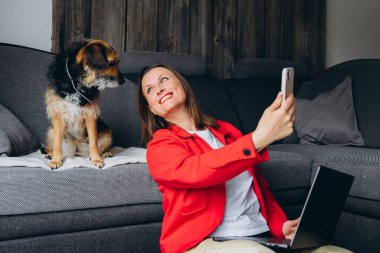 Bilgisayarla uzaktan çalışan bir kadın, evde köpeğiyle selfie çekiyor. Mutlu serbest çalışanlar evcil hayvanlarla eğleniyor, rahat yaşam alanında yerde oturuyor, gündelik dijital yaşam tarzı. Yüksek kalite fotoğraf