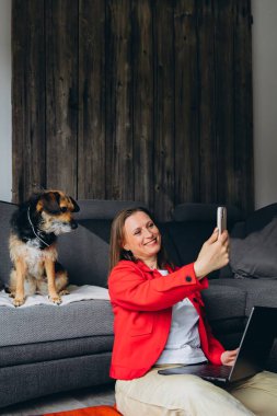 Bilgisayarla uzaktan çalışan bir kadın, evde köpeğiyle selfie çekiyor. Mutlu serbest çalışanlar evcil hayvanlarla eğleniyor, rahat yaşam alanında yerde oturuyor, gündelik dijital yaşam tarzı. Yüksek kalite fotoğraf