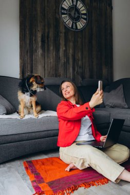 Bilgisayarla uzaktan çalışan bir kadın, evde köpeğiyle selfie çekiyor. Mutlu serbest çalışanlar evcil hayvanlarla eğleniyor, rahat yaşam alanında yerde oturuyor, gündelik dijital yaşam tarzı. Yüksek kalite fotoğraf