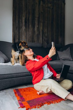 Bilgisayarla uzaktan çalışan bir kadın, evde köpeğiyle selfie çekiyor. Mutlu serbest çalışanlar evcil hayvanlarla eğleniyor, rahat yaşam alanında yerde oturuyor, gündelik dijital yaşam tarzı. Yüksek kalite fotoğraf