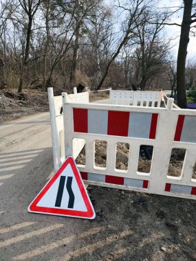 Ağaçlık bir alanda yol inşaatı. Plastik bariyerler ve şerit daraldığını gösteren bir tabela var. Kırmızı-beyaz barikata karşı düşen uyarı işareti. Erken bahar manzarası.