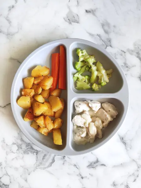 Çocuk menüsü bölünmüş bir silikon tabakta servis edilir. İçinde sotelenmiş patates, havuç çubuğu, buğulanmış brokoli ve kremalı tavuk var. Mermer zeminde yürüyen çocuklar için sağlıklı ve dengeli yiyecekler..