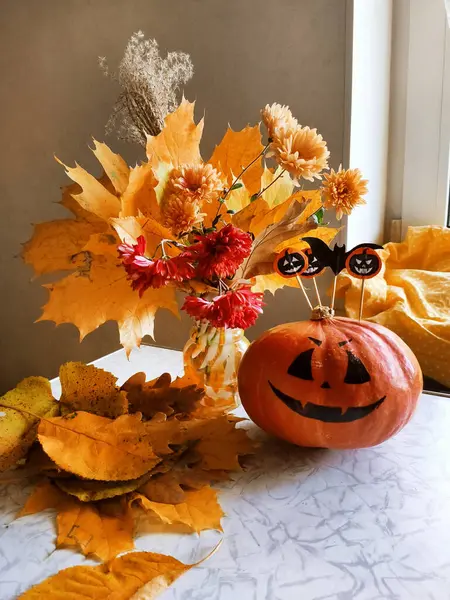 Gülen bir balkabağı ve sarı akçaağaç yapraklı bir buket portakal ve kırmızı kasımpatı ile dolu cam bir vazo. Pencerenin yanında mevsimlik kompozisyon, Cadılar Bayramı ev dekorasyonu.