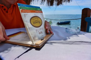 Durres, Arnavutluk - 3 Eylül 2022: Restaurant 2 Kitarrat Arome Deti. Menü kartını tutan ve okuyan, yemeği seçen adam. Güneşli bir günde Adriyatik sahilinde güzel bir restoran.