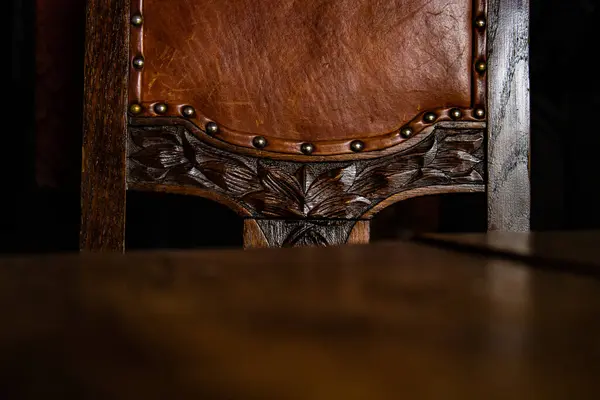 Close up of the detail of antique wooden Chair covered with brown leather upholstery, decorated with metal rivets and artistic wood carving elements. Dark background. Copy Space.