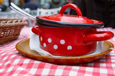 Kırmızı tencerede geleneksel lahana çorbası. Kase seramik tabakta beyaz dokularla servis edilir. Retro tarzı ekose masa örtüsü. Açık hava restoranı. Bulanık arkaplandaki insanlar.