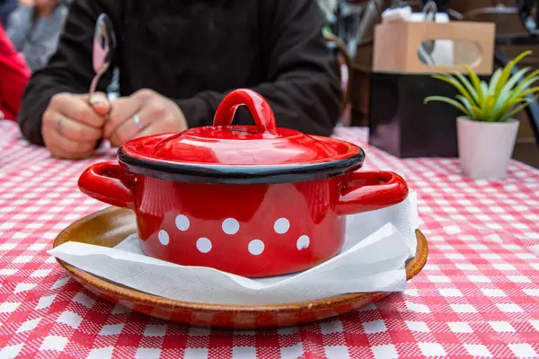 Dining out in the restaurant. Woman is holding the spoon and going to eat the traditional soup in the red cast iron pot served on the ceramic plate with white tissue. Retro style plaid table cloth.