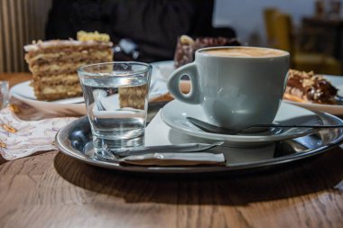 Krema köpüklü bir fincan kahve kaşıkla beyaz tabakta servis edilir. İçinde kek ve tatlı olan birkaç tabak, bir bardak su, kahve dükkanındaki ahşap sehpanın üzerindeki metal tepside şeker. Seçici odak