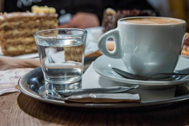 Krema köpüklü bir fincan kahve kaşıkla beyaz tabakta servis edilir. İçinde kek ve tatlı olan birkaç tabak, bir bardak su, kahve dükkanındaki ahşap sehpanın üzerindeki metal tepside şeker. Seçici odak