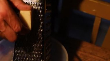 Adam parmesan peynirini ahşap bir masada plastik bir haykırışla ovuyor. Metal rendeleyiciye peynir sürterler. Peynirli yemek pişiriyorum. Akşamları mutfakta çalışıyorum. Koyu arkaplan.