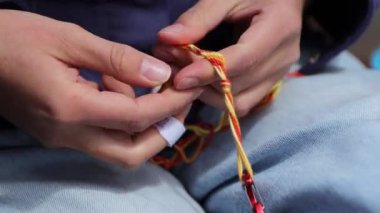 Sarı turuncu saç örgülü arkadaş bileziği ya da kemeri yapan genç kıza yakın çekim. Pamuk ipliklerini ören dişilerin elleri üzerlerinden geçerken çok güzel desenler oluşturuyor. El yapımı konsept.
