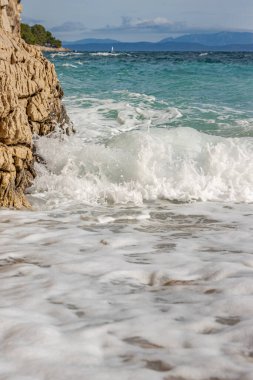 Kayalık bir plaja çarpan görkemli yumuşak dalgalar. Dalga taşlı bir kıyıda kırılarak bir sprey oluşturur. Manzaralı deniz manzarası. Kart, afiş, seyahat katalogu fotoğrafı. Uzayı kopyala Seçici odak.