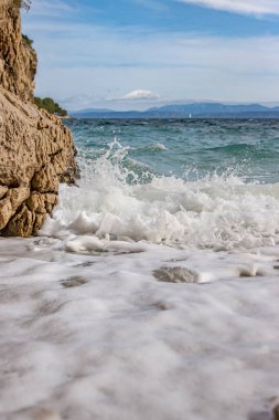 Kayalık bir plaja çarpan görkemli yumuşak dalgalar. Dalga taşlı bir kıyıda kırılarak bir sprey oluşturur. Manzaralı deniz manzarası. Kart, afiş, seyahat katalogu fotoğrafı. Uzayı kopyala Seçici odak.